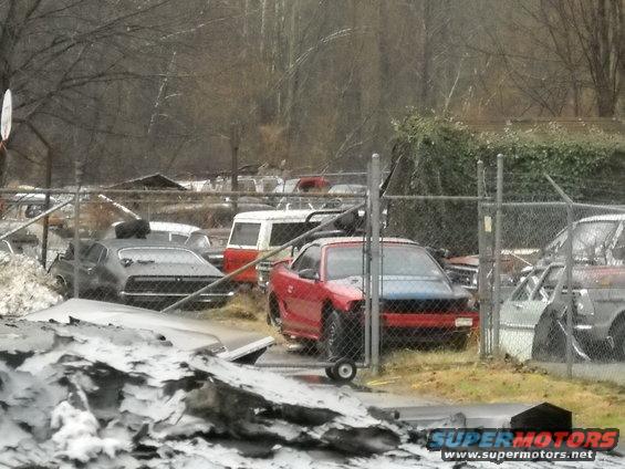 Junkyard scene with a red car, a grey car, and a white van visible behind a fence