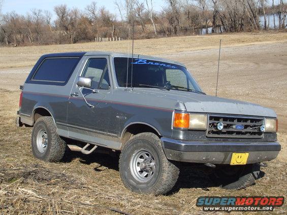 Gray and dark gray Ford Bronco with a blue 'Bronco' decal on the windshield, parked on dry grass
