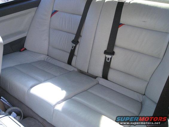 Light gray leather rear car seats with black seatbelts and a red, white, and blue stripe detail on the headrest