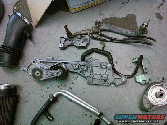 Engine parts laid out on a concrete floor, including an air intake tube, a tensioner assembly, and hoses