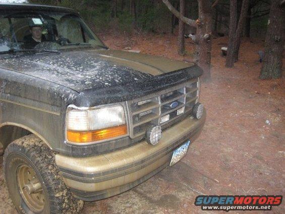 Front end of a dirty Ford Bronco parked in a wooded area. A person is visible in the driver's seat