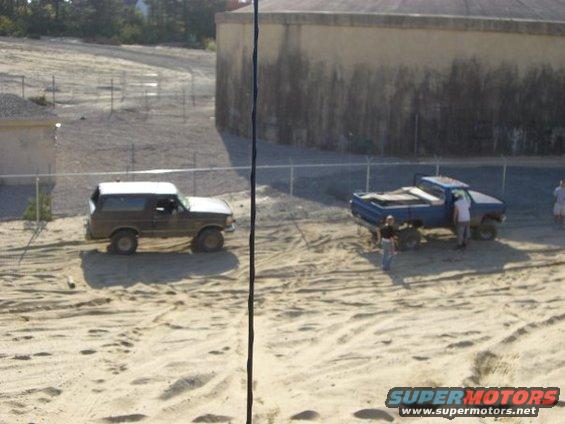 Brown Bronco and blue pickup truck on sandy terrain near a large concrete structure