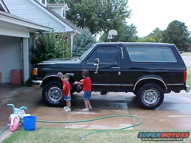 Black Ford Bronco parked in driveway with two children washing it. A tricycle and bucket are nearby