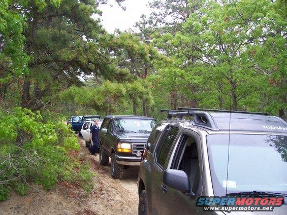 Line of SUVs driving on a dirt road through trees. A person in a blue shirt stands by a dark SUV