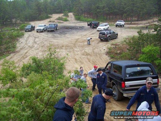 Black Ford Bronco from rear, with silver stripe. People stand around it on a dirt track