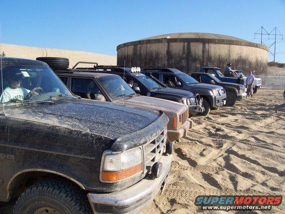 Several SUVs and trucks parked in sand dunes, with a large concrete structure in the background