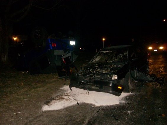 Two wrecked cars on a dark, wet road at night. One car is black with its hood open and front end damaged. Another blue vehicle is overturned nearby