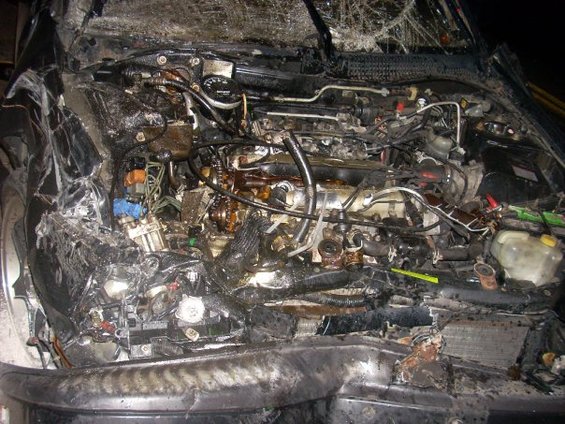 Damaged engine compartment of a dark-colored vehicle, showing extensive fire and impact damage