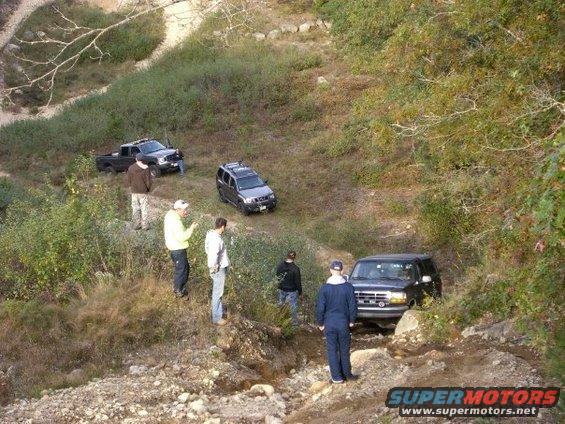 Several people watch a black 1994 Ford Bronco navigate a rocky off-road path