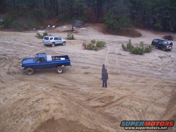 Blue pickup truck, silver SUV, and black SUV on a sandy track with trees in the background