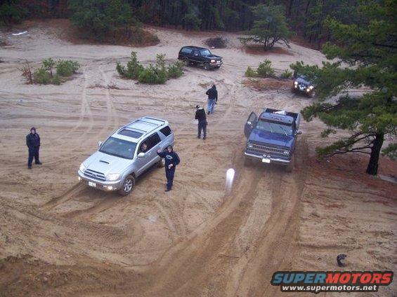 Several SUVs and a pickup truck are parked and driving on a sandy, wooded trail. People stand around the vehicles