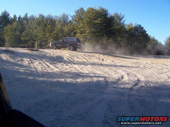 1994 Ford Bronco driving on a dirt road, kicking up dust. Trees and blue sky in background