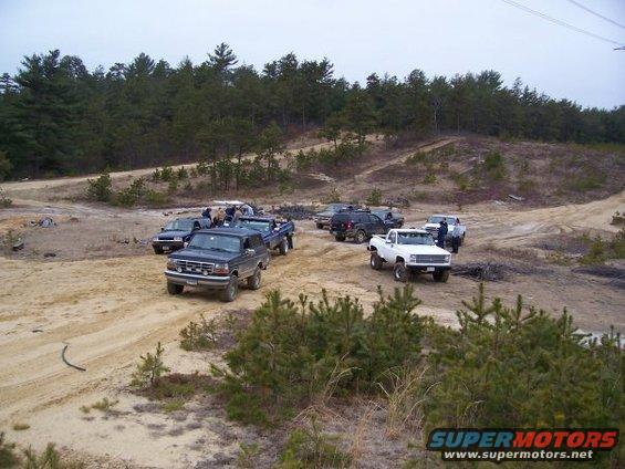 Several trucks and SUVs parked on a dirt track in a wooded area. One dark blue SUV is in the foreground