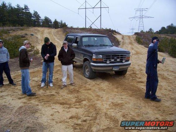 Dark blue Ford Bronco parked on a dirt hill with a group of guys standing around it. Power lines visible in the background