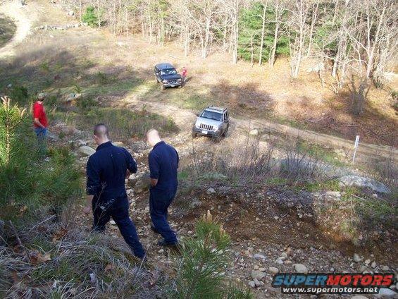 Two SUVs on a dirt road in a wooded area, with people nearby
