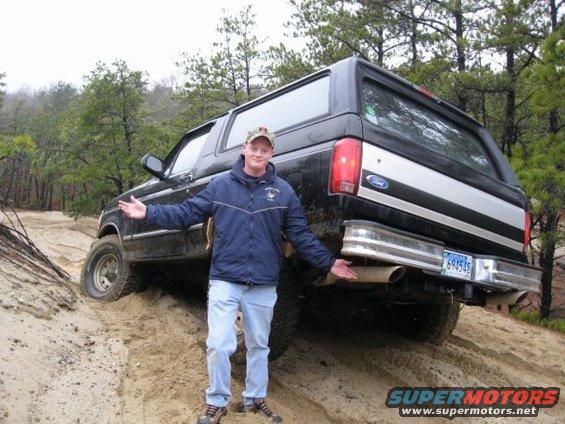 Black Ford Bronco with a man standing in front, angled rear view, on a sandy incline