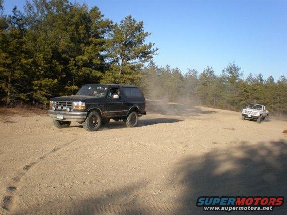 Black 1992 Ford Bronco driving on a dirt road, kicking up dust. Another white truck is visible in the background