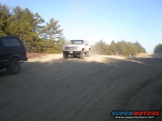 White pickup truck driving on dirt road, black SUV parked to the left