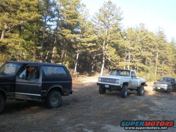 Black SUV, silver pickup truck, and dark pickup truck parked on dirt road with trees in background