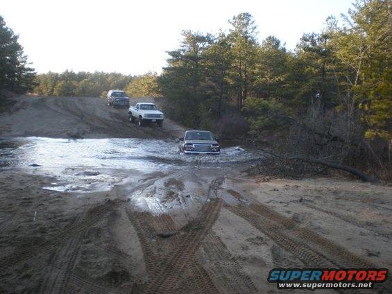 three trucks driving through a muddy, watery path in a wooded area