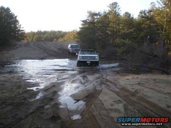 White 1987 Ford Bronco driving through water on a dirt road, with another vehicle behind it