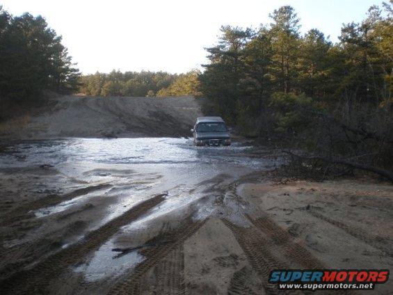 Dark SUV driving through water on a dirt road with trees in background
