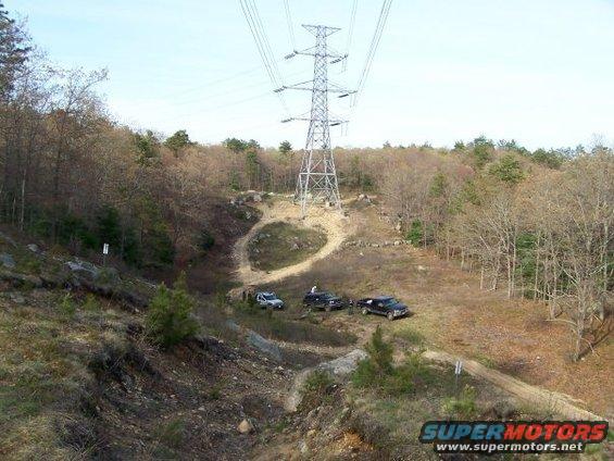 Three SUVs parked on a dirt path below a large power line tower in a wooded area
