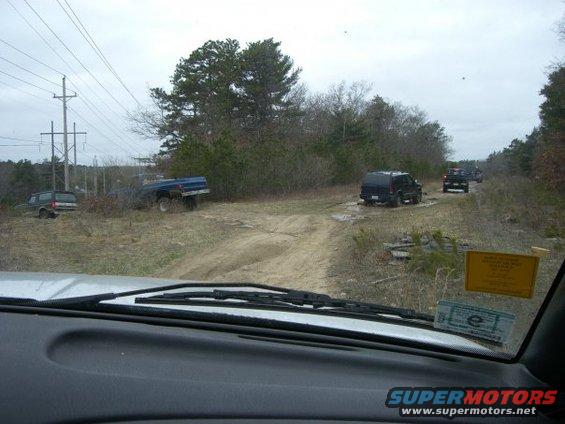 Several SUVs and a blue pickup truck are parked and driving on a dirt road with trees and power lines