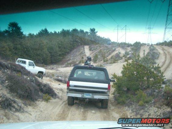 Black and silver Ford Bronco driving on a dirt road, rear view. License plate is visible