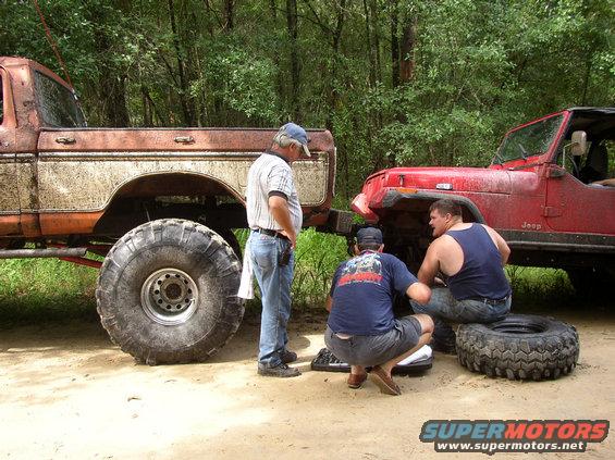 Men working on a red Jeep next to a lifted, muddy truck