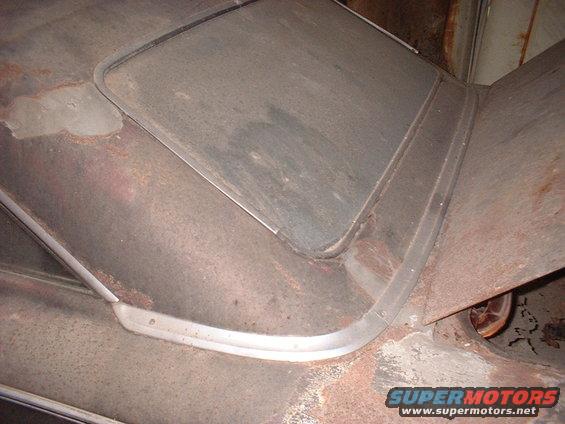 Close-up of a rusty car roof with a metal trim around a window opening. A wheel is visible on the right