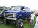 1986 Ford Bronco - A lifted blue GMC van with massive tires is displayed at an outdoor event, with another vehicle visible to the left.
