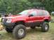 1986 Ford Bronco - A lifted red Jeep Grand Cherokee is parked on grass with trees in the background.