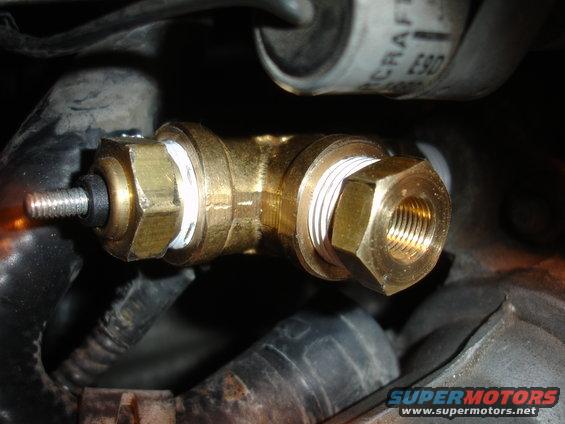 Brass T-fitting with threaded ends and a sensor attached to one side, surrounded by dark engine components