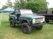 1986 Ford Bronco - A teal Ford Bronco is parked on a grassy field next to a black pickup truck under a blue tent.