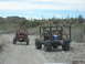 1996 Ford Bronco - Two custom off-road vehicles navigate a dusty desert trail under a bright blue sky with saguaro cacti in the background.