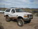 1996 Ford Bronco - This lifted white Ford Bronco is parked in a dusty, outdoor area with other vehicles visible in the distance.