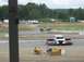 1986 Ford Bronco - A car is captured mid-air as it jumps a dirt ramp during an event, with spectators watching.