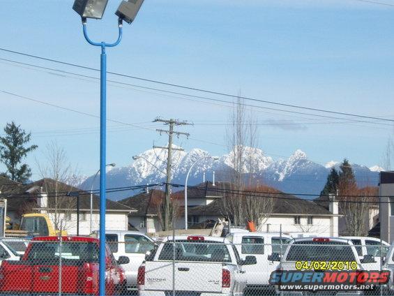 Red pickup truck and white pickup trucks behind a chain-link fence with snowy mountains in the background