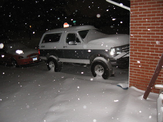 Two-tone Ford Bronco parked in driveway during snowfall, red car behind it