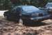 1987 Ford Thunderbird - A dark-colored Ford Thunderbird coupe is seen from the rear, parked on a dirt lot with trees in the background.