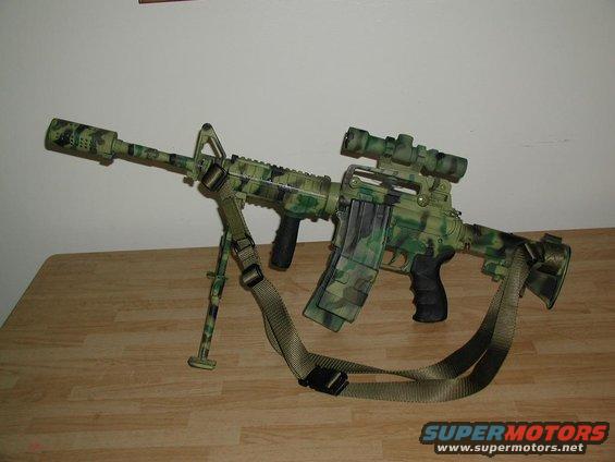 Camouflaged assault rifle with bipod, scope, and sling on a wooden table