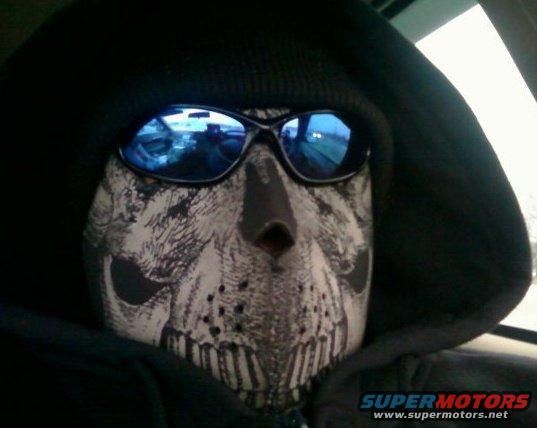 Person in skull mask and sunglasses, reflection shows dashboard and road