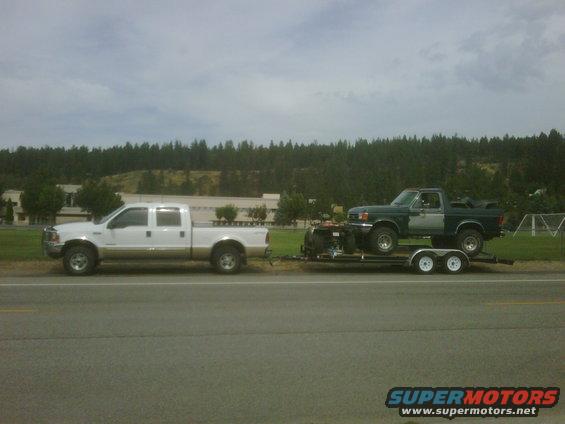 White crew cab pickup truck towing a trailer with a green pickup truck on it
