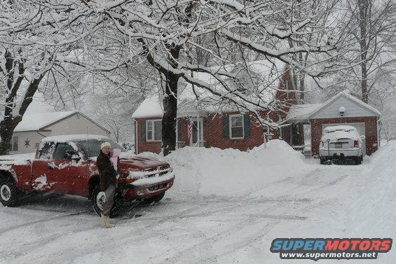 Person holding baby next to red pickup truck covered in snow, house and another vehicle in background