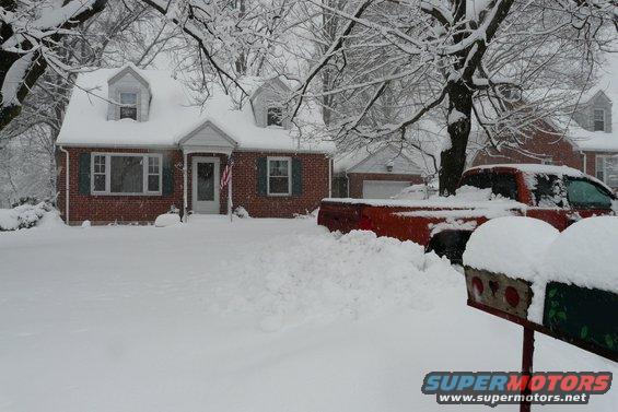 Red pickup truck partially buried in snow, parked in front of a brick house during a snowstorm