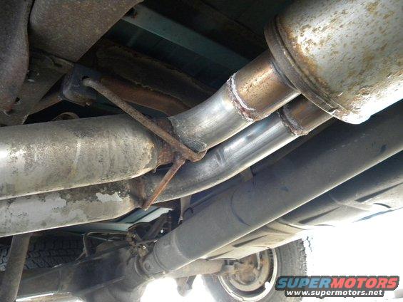Underside view of a vehicle's exhaust pipes, muffler, driveshaft, and rear tire