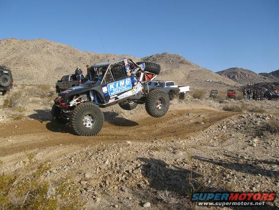 Buggy airborne over dirt track, desert mountains in background. Another truck and buggy visible