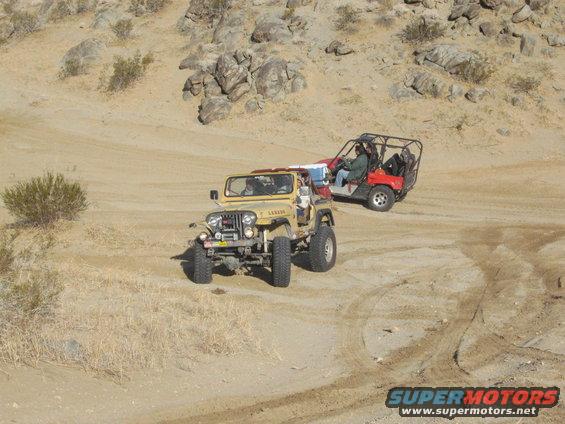tan Jeep CJ-7 driving on a dirt trail with a red dune buggy behind it