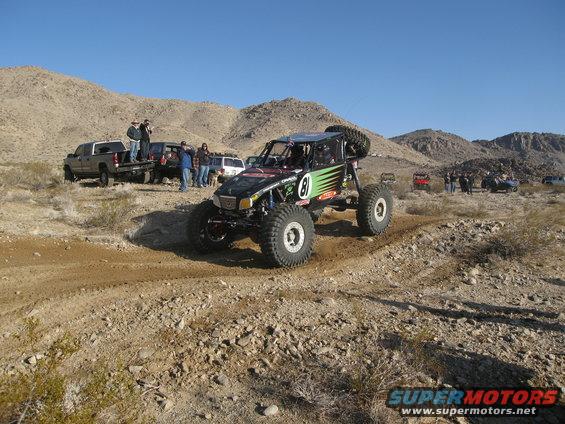 Custom off-road buggy with number 81 on side, driving on dirt track in desert landscape. Other trucks and people in background